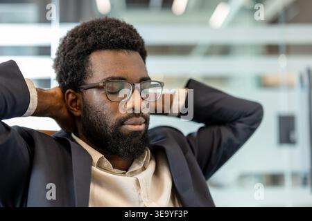 Executive man with afro hair and eyeglasses finding relief from stress, leaning back with hands behind head and eyes closed, enjoying a moment of calm relaxation in the corporate office environment Stock Photo
