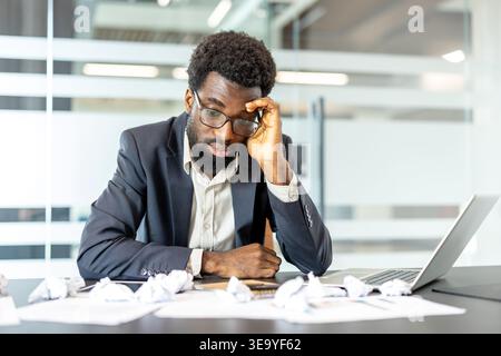 Male professional experiencing burnout and frustration while working at a desk, surrounded by crumpled papers and using a laptop, coping with workplace pressure and professional challenges Stock Photo
