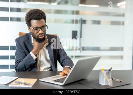 Young businessman suffering from a sore throat and discomfort while using a laptop at his office desk, indicating illness and struggling to maintain productivity Stock Photo
