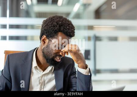 Professional man feeling mental pressure and discomfort, closing eyes and pressing his fingers to his forehead in an office, symbolizing corporate burnout and the need for relief Stock Photo