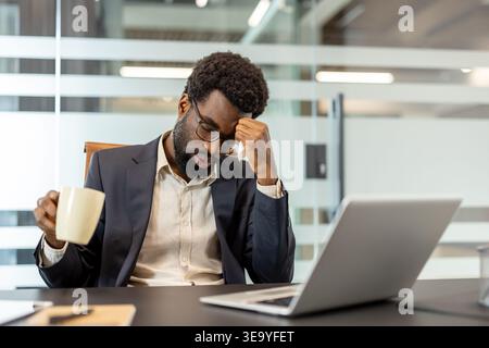Young man feeling unwell and struggling with a headache while holding a coffee cup, sitting at his office desk with a laptop, experiencing stress and sickness during a demanding workday Stock Photo