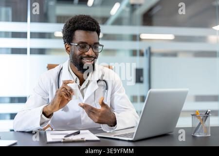 Male doctor wearing a lab coat and stethoscope engaging in a remote video call on a laptop, discussing medication by showing a prescription bottle to the screen in a modern office Stock Photo
