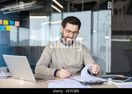 Businessman working with paperwork and financial data, reviewing documents on his desk with a laptop and calculator, demonstrating focus and productivity in a contemporary workspace Stock Photo