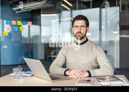 Young businessman sitting at desk looking at camera while working on laptop in a contemporary office setting, representing technology, ambition, and a professional workspace Stock Photo