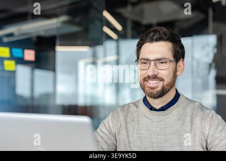Happy professional man wearing casual sweater and eyeglasses smiling while using a laptop, representing success, productivity, and positive working environment Stock Photo