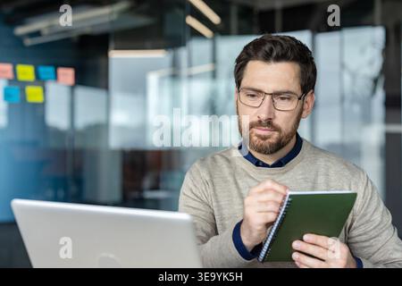 Confident bearded man with eyeglasses focused on work, using a laptop and writing in a notebook, concentrating on planning tasks and improving productivity in a modern office environment Stock Photo