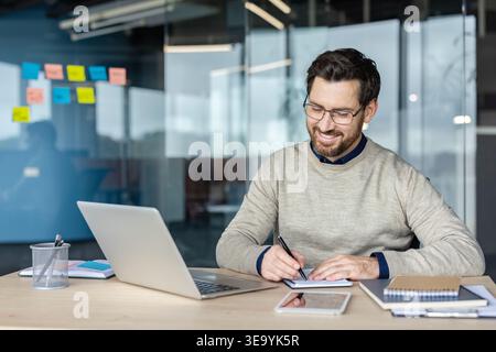 Smiling man wearing glasses working at his office desk, using a laptop and writing notes in a notebook, with sticky notes on a glass wall in the background Stock Photo