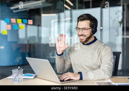 Businessman wearing a headset with microphone using a laptop for virtual communication, participating in a remote meeting, or teleconferencing from his modern workplace desk Stock Photo