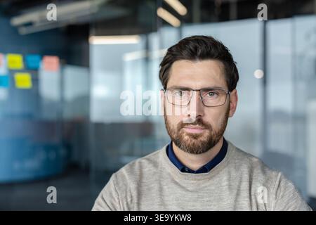 Confident bearded professional man wearing eyeglasses and a crewneck sweater, standing in a modern office with glass partitions, portraying leadership and corporate success Stock Photo