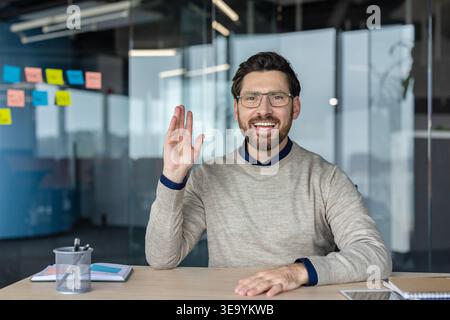 Smiling businessman wearing eyeglasses waving hand communicating online from modern office, looking at camera and participating in a video conference or virtual interview Stock Photo