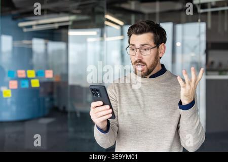 Man in a modern office looking surprised and frustrated after reading upsetting information or urgent message on his mobile phone, showing a strong emotional reaction Stock Photo