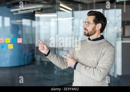 Confident businessman wearing glasses pointing fingers at colorful sticky notes on a clear glass board, strategizing during a meeting in the bright, contemporary office workspace Stock Photo