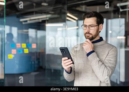 Businessman with a thoughtful expression checking his mobile phone in a contemporary office setting, reflecting on communication, technology, and business solutions Stock Photo