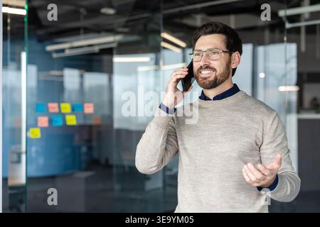 Happy bearded man with glasses discussing on a mobile phone, actively communicating and expressing ideas while working in a contemporary office environment Stock Photo