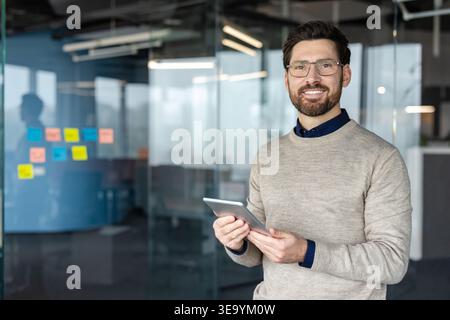 Professional businessman with beard and glasses holding a digital tablet, standing confidently in a modern glass office, representing technology, communication, and connectivity in business Stock Photo