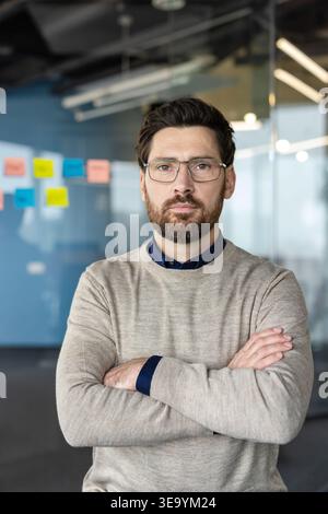 Serious bearded professional wearing glasses stands with arms crossed in a modern glass-walled office, confident entrepreneur focused on strategy and leadership with sticky notes behind him Stock Photo