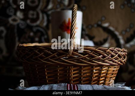 A round wicker basket with a braided handle stands before a floral mug in subdued light. The artisan weave and warm wood tones shape a nostalgic Stock Photo