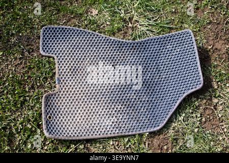A used rubber car mat lies on patchy grass after washing. Honeycomb texture and scuffs reveal hard service while bright sunlight and top down framing Stock Photo