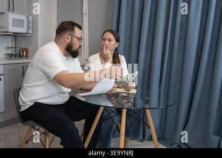 Young couple sitting at a glass table in a modern kitchen reviewing documents and calculating money, feeling stress and worry while discussing househo Stock Photo