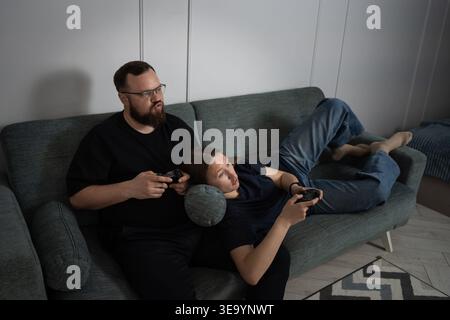 Young couple on a sofa at home, focused and smiling while playing a competitive console video game with wireless controllers, enjoying relaxed weekend Stock Photo