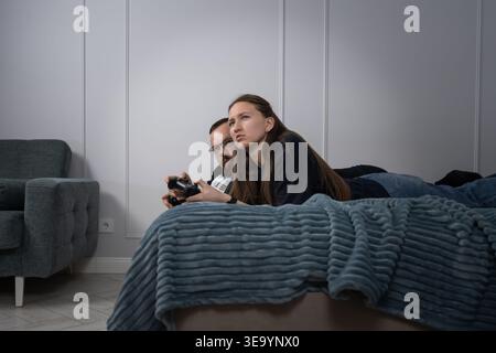 Young couple lying on a bed with a blue blanket, intensely concentrating on their video game as they use controllers for competitive gaming in a moder Stock Photo