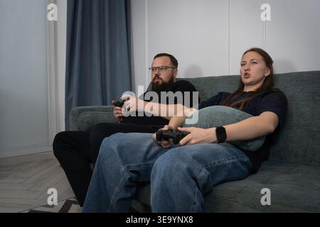 Young couple enjoying leisure time at home, concentrating on their game controllers while sitting on a comfortable sofa and engaging in friendly compe Stock Photo