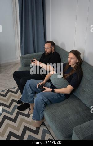 Man and woman concentrating on playing video games with controllers while sitting on a comfortable sofa in their modern living room, enjoying entertai Stock Photo