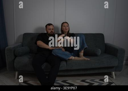 Young couple cuddled on a comfortable sofa in a dim living room, enjoying a cozy movie night with popcorn and snacks, relaxed and spending quality tim Stock Photo