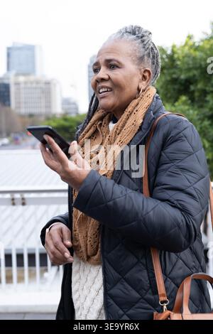 Vertical video: African American Senior woman on bridge, using phone assistant, holding brown bag Stock Photo