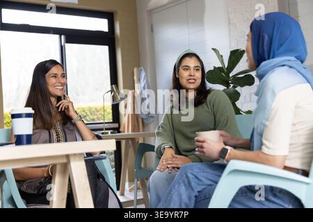 Three women colleagues chatting around wood table in office, wearing headscarf and holding mug Stock Photo