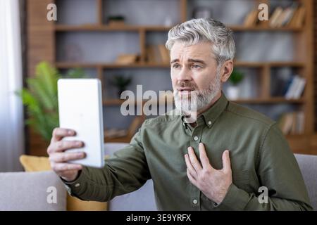 Senior man having a video consultation with a doctor using a tablet, expressing discomfort and gesturing towards his chest, symbolizing telemedical health challenges and digital healthcare support Stock Photo