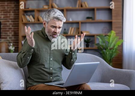 Mature man experiencing digital detox and technology issues, looking surprised and shocked at his laptop screen with hands raised while sitting on a sofa at home Stock Photo