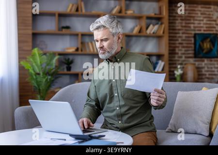 Mature man sitting on a sofa, focused on a laptop and holding paper documents, managing personal finances and doing online banking in a comfortable living room environment Stock Photo