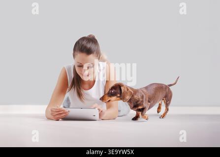 Young woman lying on floor using tablet while curious dachshund dog looks at screen. Home lifestyle, technology use and pet companionship concept on n Stock Photo