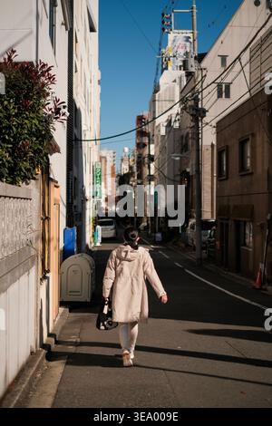 Woman walks down a sunlit narrow urban street with buildings and utility poles Stock Photo