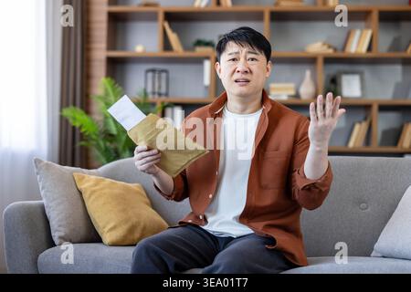 Young asian man sitting on a sofa in his living room, opening an official envelope and showing a confused expression and stop gesture, receiving unexpected bad news, debt, or tax notification Stock Photo