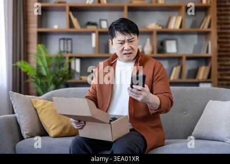 Asian man sitting on a sofa. Looking at a smartphone with a frustrated expression while holding an open parcel. Dealing with a poor unboxing experience. Customer complaint. Or failed delivery at home Stock Photo