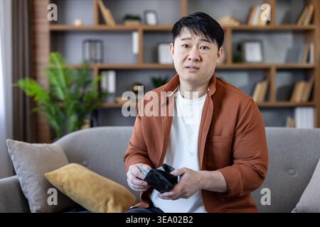 Man feeling sad and stressed, realizing bankruptcy and financial problems while holding an empty wallet, sitting on a sofa in a modern living room setting Stock Photo