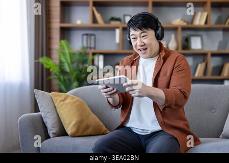 Excited asian man wearing headphones enjoying an exciting mobile game on a tablet while relaxing on the couch in his modern living room at home, finding leisure and entertainment Stock Photo