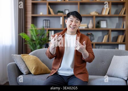 Smiling asian man sitting on a comfortable modern couch in a living room, actively gesturing while talking, representing communication, discussion, and remote work concepts Stock Photo