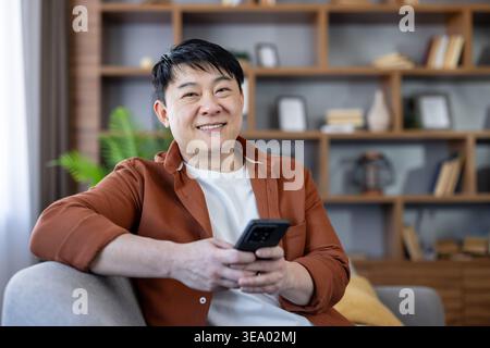 Smiling asian man sitting comfortably on a sofa in his living room, holding a smartphone and looking at the camera, symbolizing relaxation and digital connection at home Stock Photo
