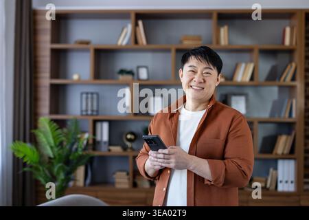 Happy asian man using a smartphone and smiling, standing in his modern home office with a bookshelf in the background, representing remote work and connectivity Stock Photo