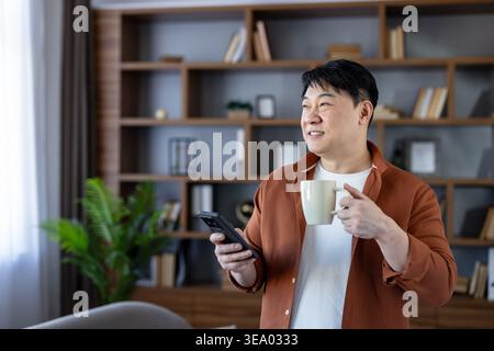 Cheerful asian man holding a coffee mug and smartphone, looking away with a smile, standing in a smart casual outfit in a modern home office with a bookshelf background Stock Photo
