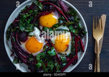 A nutritious breakfast: three fried eggs placed on sauteed beet greens and stems on a white plate with gold cutlery. High-protein vegetarian keto diet Stock Photo