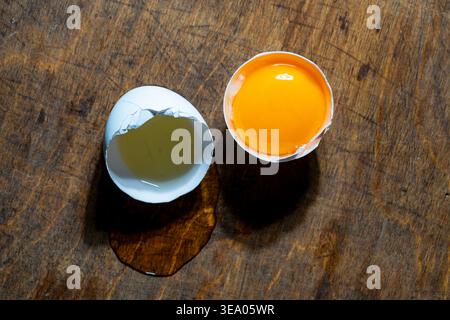 Fresh cracked egg with bright yolk. Raw chicken egg broken on dark wood background. One shell holds a vibrant orange yolk while egg white spills onto Stock Photo