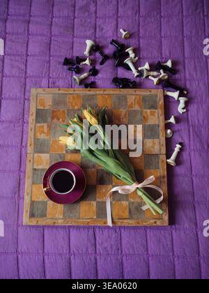 Vintage Porcelain Coffee Cup and Yellow Tulips on Rustic Wooden Chessboard with Scattered Chess Pieces on Purple Textile — Romantic Morning Flat Lay Stock Photo