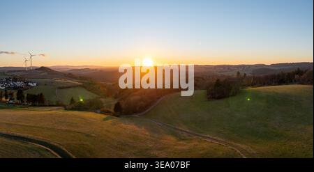 Aerial view of the sun setting behind wind turbines casting long shadows over rolling green fields, Hellenthal, Germany. Stock Photo