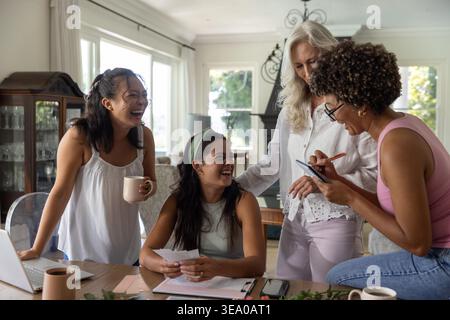 Diverse female friends reviewing papers and using laptop at wooden dining table with mugs. Teamwork, collaboration, home, warm, bright, multigenerational, tablet Stock Photo