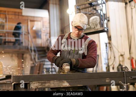 Young man in overalls and safety glasses using angle grinder creating sparks while working on steel frame Stock Photo