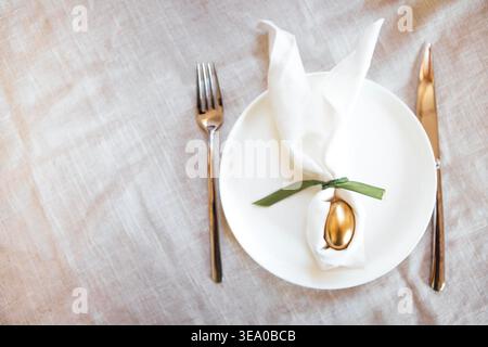 Elegant table arrangement with a white plate displaying a golden egg wrapped in a napkin, flanked by a fork and knife on a linen tablecloth Stock Photo